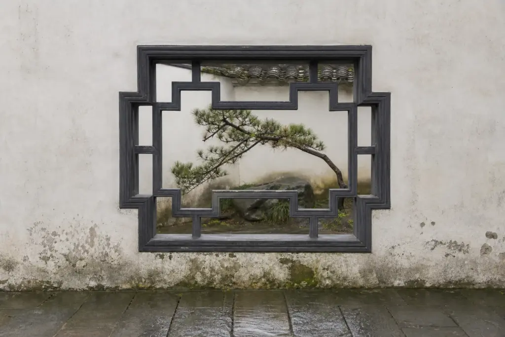 Geometric lattice window in a white garden wall framing a pine branch and small stone courtyard.