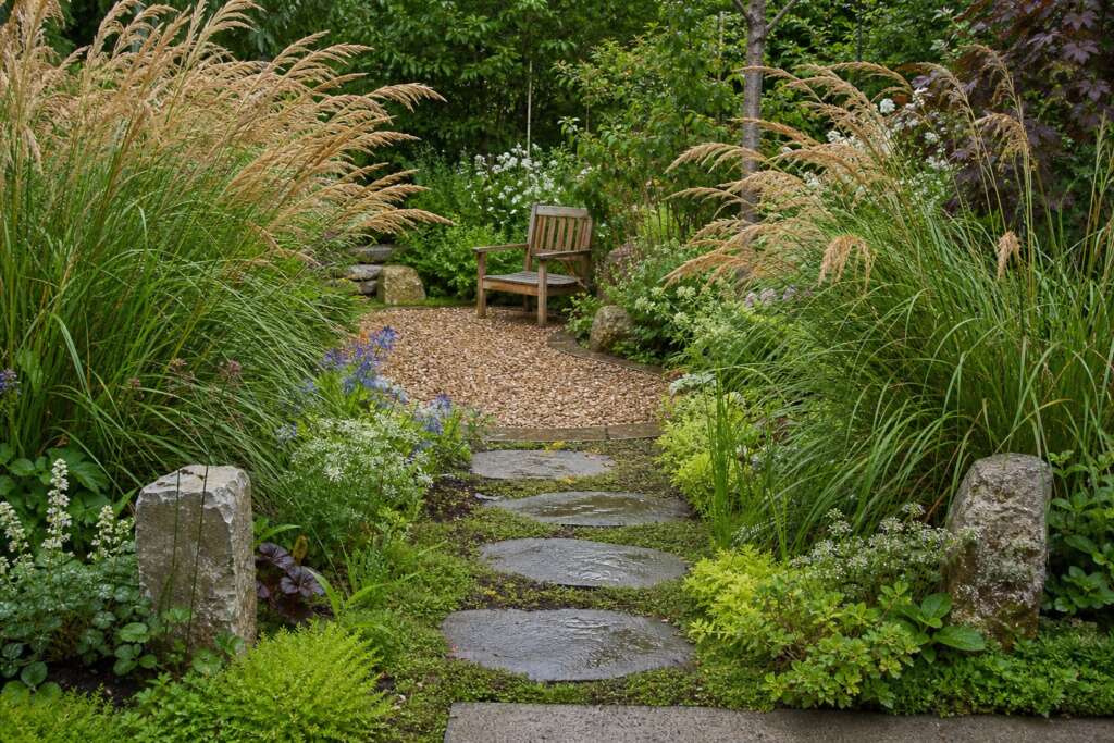 A realistic garden threshold with tall grasses, stepping stones, gravel, and a wooden bench beyond.