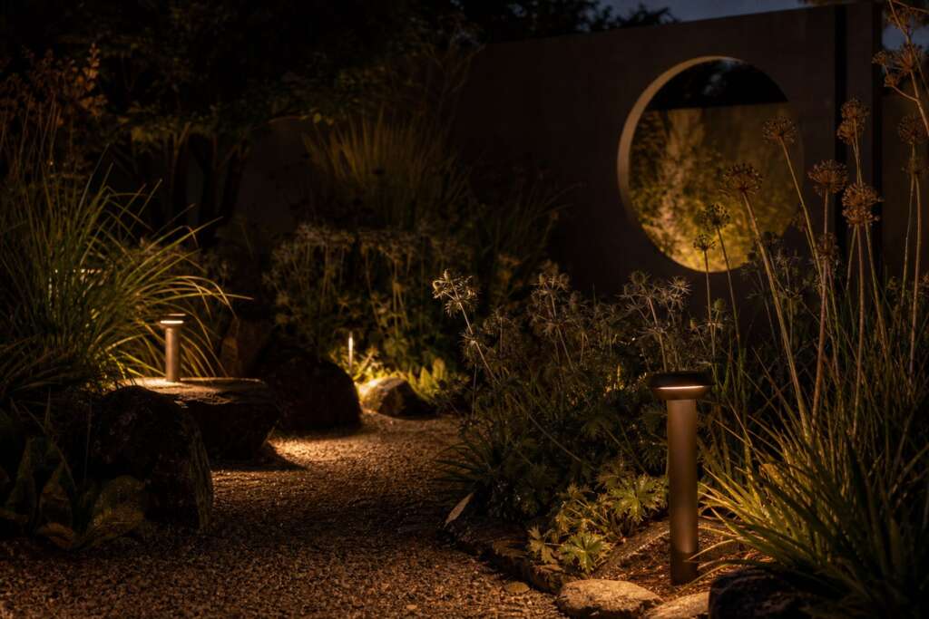 Low shielded bollard lights illuminate a gravel path, grasses, seed heads, and a dark concrete screen with a circular opening at night.